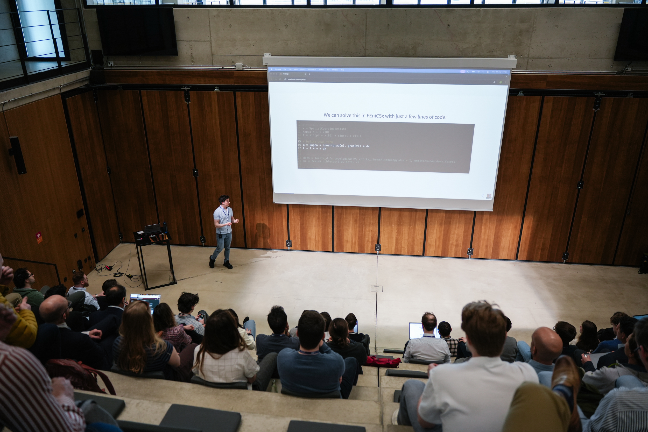 A speaker presents FEniCSx code to a full lecture hall at OSSFE 2026